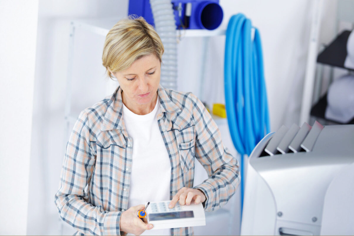 Blond woman checking the thermostat for her home HVAC system. She's holding a screwdriver and the thermostat in her right hand, pressing a button on the device with her left.