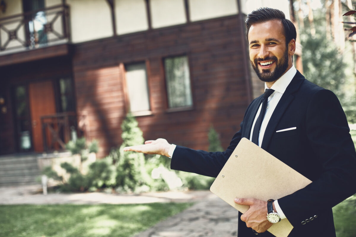 Smiling realtor holding a clipboard, gesturing toward a house.