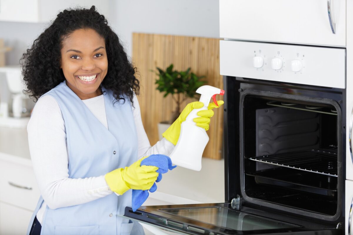 Smiling woman wearing yellow gloves, holding spray bottle and cloth, cleaning an oven.